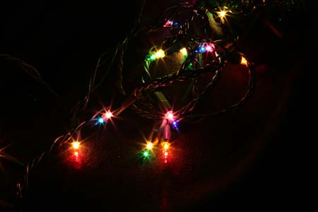 Shows lit Christmas lights on a string, in-focus with beams of light coming from the lights.